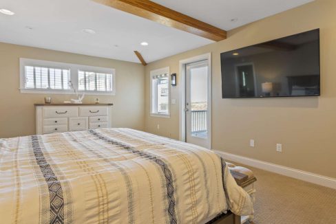 A bright and cozy bedroom featuring a neatly made bed, a dresser under a window, exposed ceiling beams, and a wall-mounted flat-screen tv.