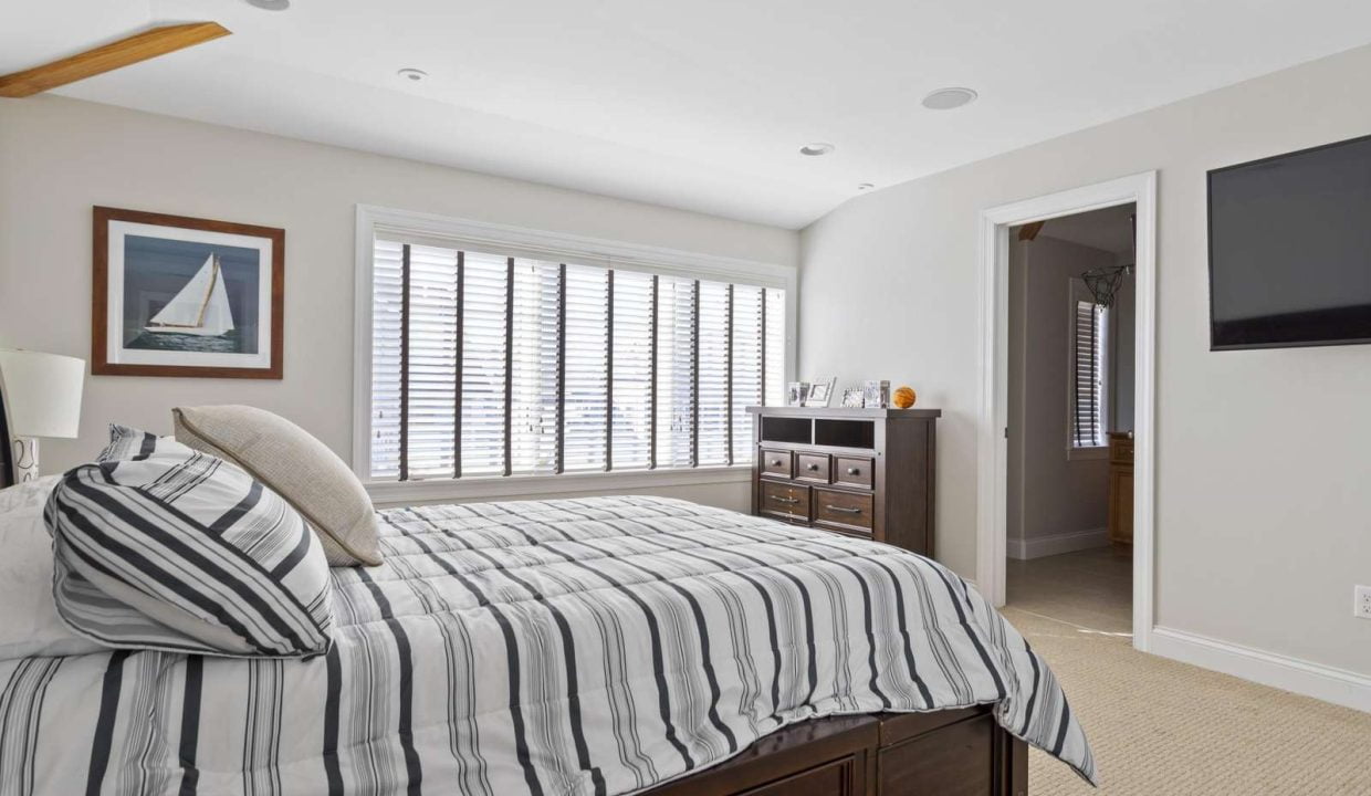 A bright, well-lit bedroom with a neatly made bed, striped bedding, plantation shutters on the windows, and a flat-screen tv on the wall.