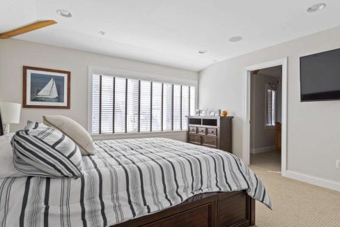 A bright, well-lit bedroom with a neatly made bed, striped bedding, plantation shutters on the windows, and a flat-screen tv on the wall.