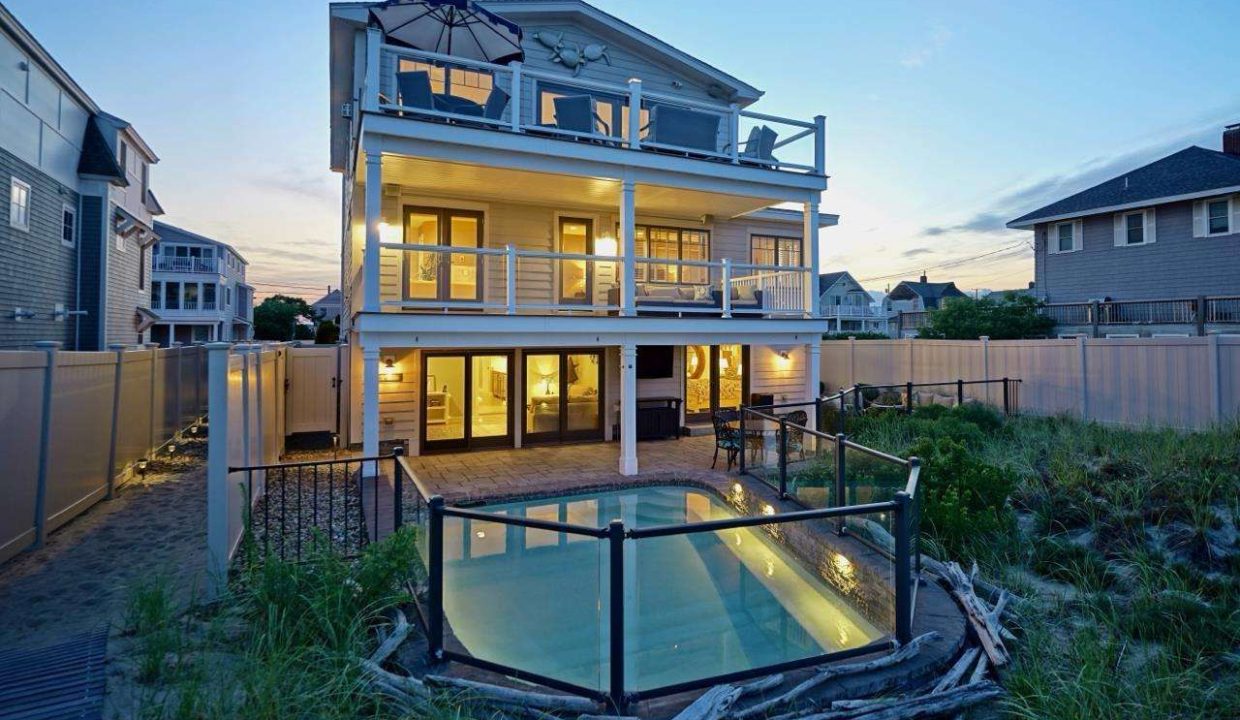 Modern multi-story beach house illuminated at dusk with surrounding fencing and dune grass.