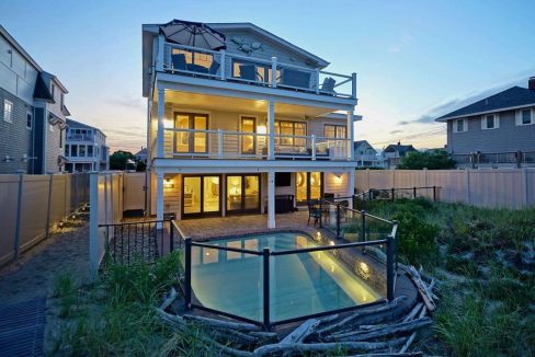 Modern multi-story beach house illuminated at dusk with surrounding fencing and dune grass.