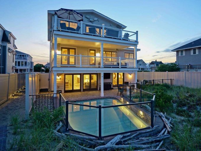 Modern multi-story beach house illuminated at dusk with surrounding fencing and dune grass.