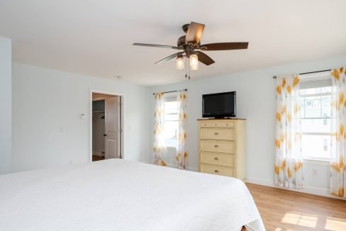 A bright bedroom with a ceiling fan, a dresser, a mounted tv, and yellow patterned curtains.