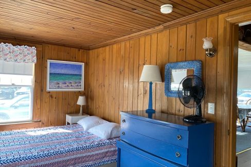 A cozy bedroom with wooden walls, a blue dresser, a patterned bedspread, and a seascape painting.