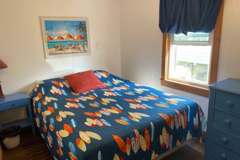 A tidy bedroom with a single bed featuring a colorful surfboard-themed blanket, a beach scene painting above the headboard, and coordinating blue furniture.