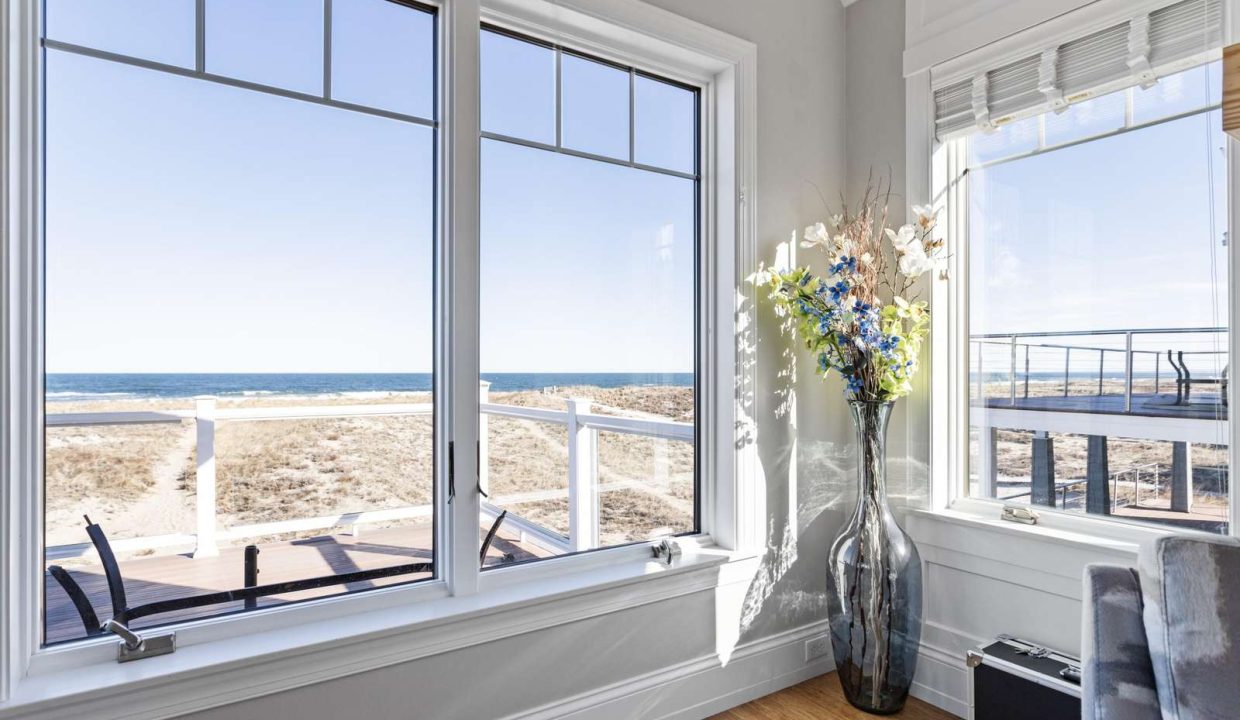 A bright, serene corner room with large windows offering a view of the beach and a vase of flowers on the windowsill.