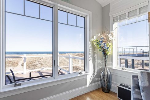 A bright, serene corner room with large windows offering a view of the beach and a vase of flowers on the windowsill.