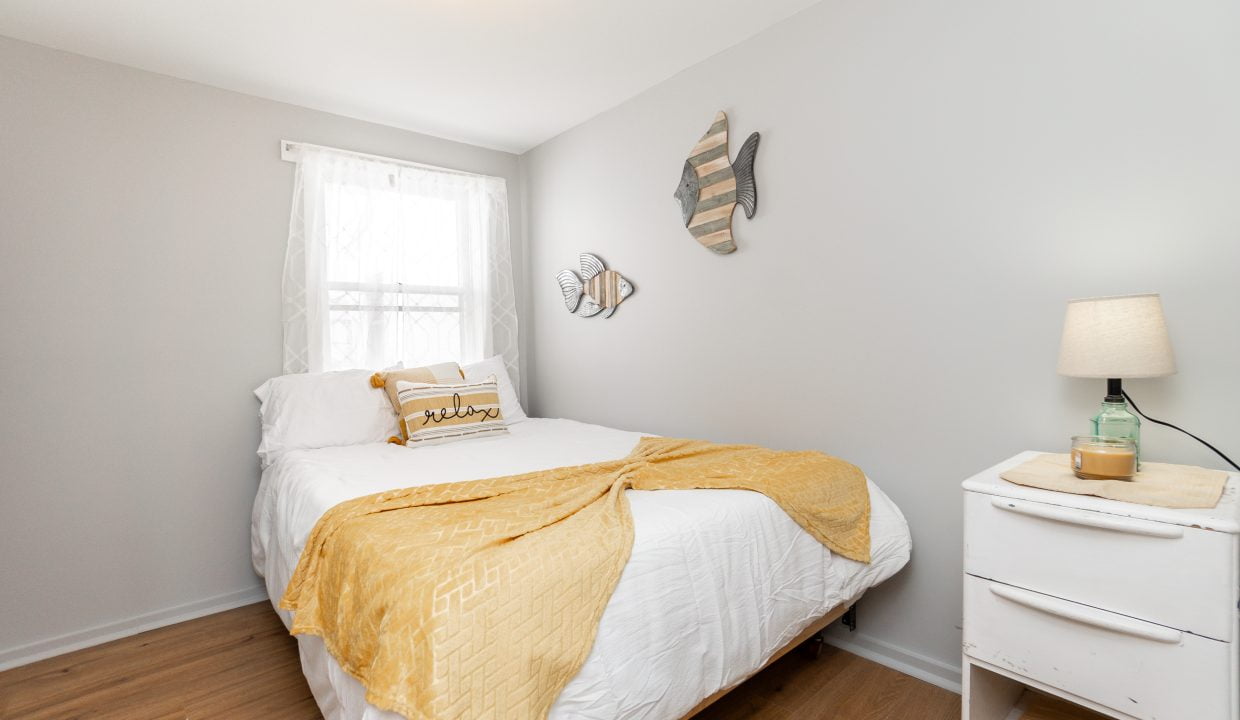 A neat and simplistic bedroom with white bedding, a yellow throw blanket, a bedside table with a lamp, and decorative wall items.