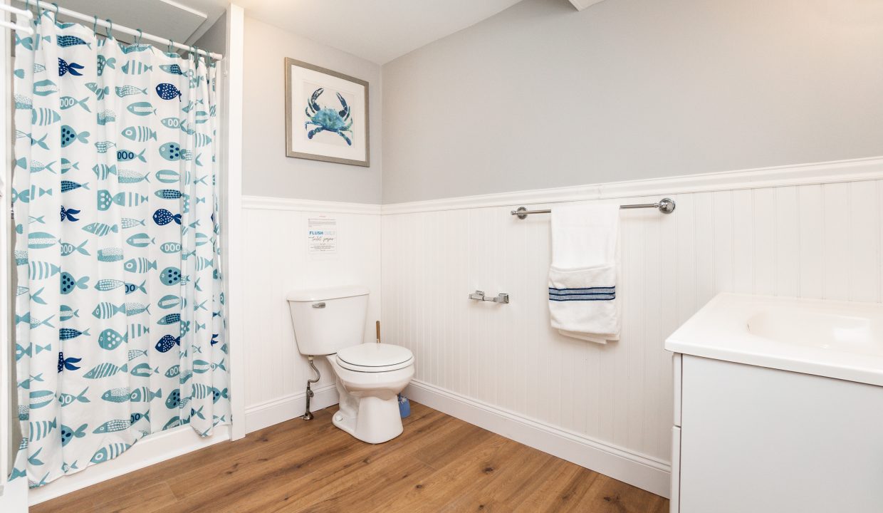 A clean and bright bathroom with a fish-themed shower curtain, white fixtures, and wood flooring.