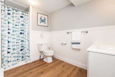 A clean and bright bathroom with a fish-themed shower curtain, white fixtures, and wood flooring.