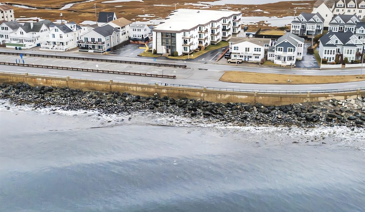 Aerial view of a coastal neighborhood with modern houses and apartments alongside a road and rocky shoreline.