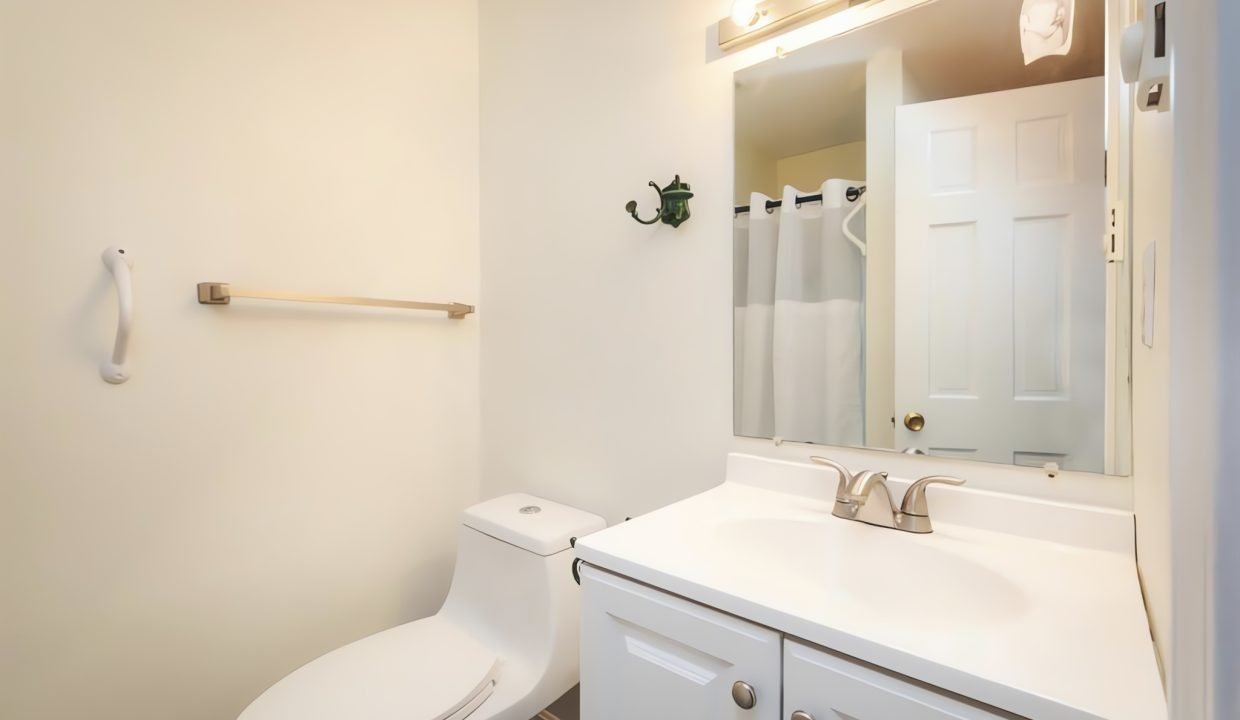 A small, well-lit bathroom with a white toilet, sink cabinet, mirror, and a towel on the rack.