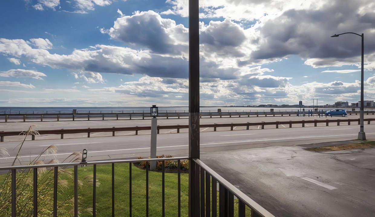 View from a balcony overlooking a sunlit road and ocean with fluffy clouds above.