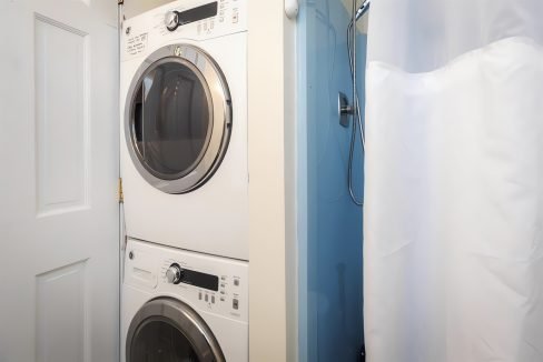 A modern laundry room featuring a stacked white washer and dryer beside a blue door and a white shower curtain.