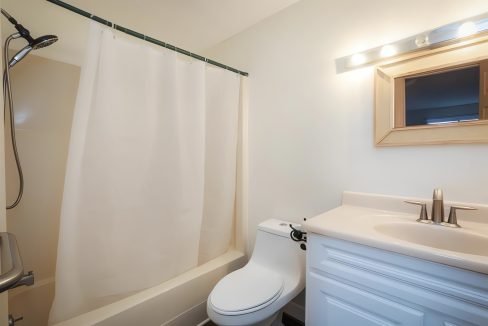 A small, well-lit bathroom with a white shower curtain, toilet, and sink vanity under a mirrored cabinet.
