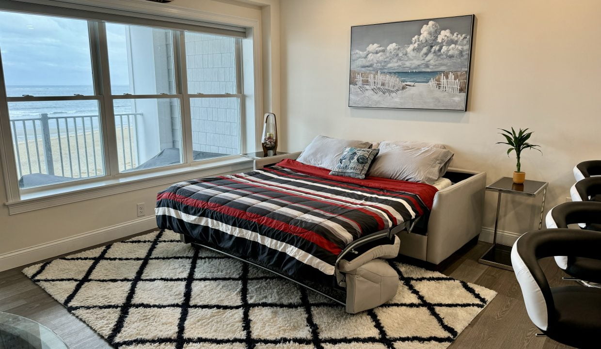 A neatly arranged bedroom with a striped comforter, a harbor-themed artwork, and a view of the water through large windows.