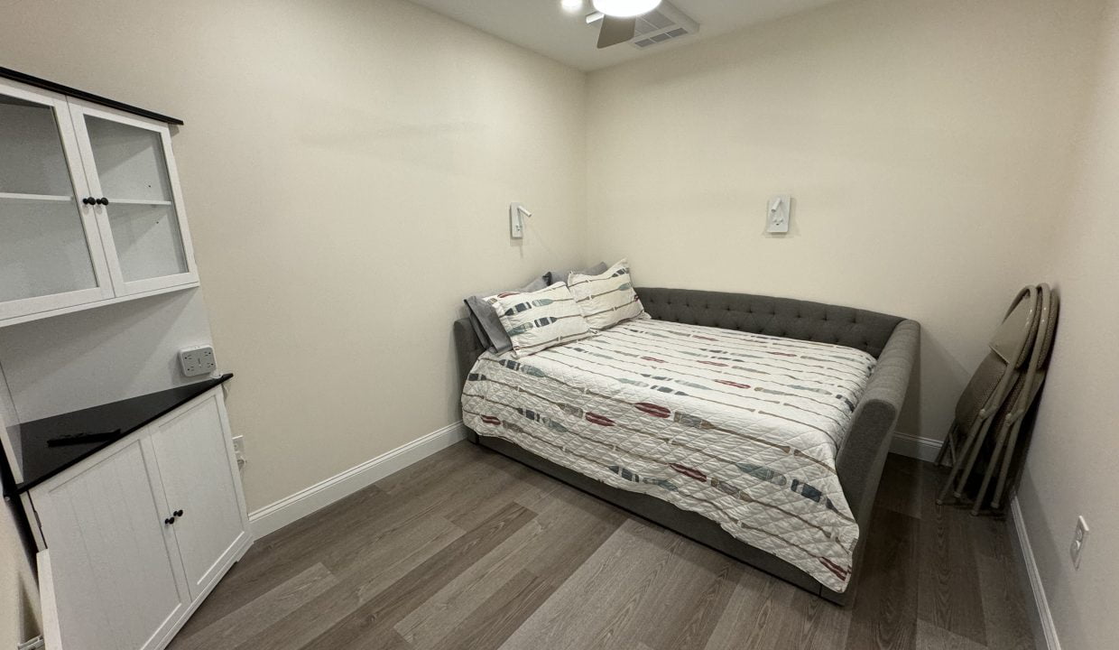 A small, well-lit room with a single bed, a ceiling fan, a black cabinet, and a mirror leaning against the wall.