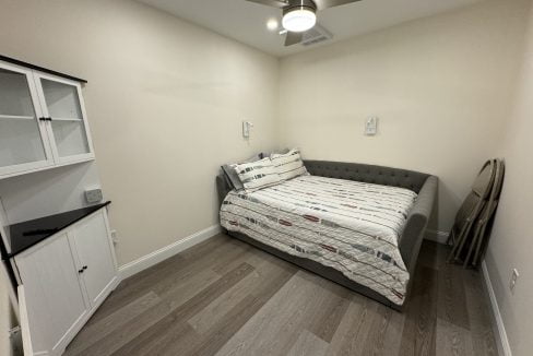 A small, well-lit room with a single bed, a ceiling fan, a black cabinet, and a mirror leaning against the wall.