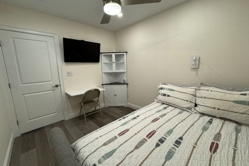 A small, neat bedroom with a bed covered in a patterned quilt, a wall-mounted tv, a desk with a chair, bookshelf, and a ceiling fan.