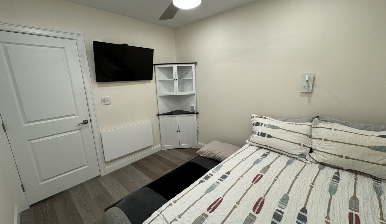 A neatly arranged, small bedroom with a bed, mounted television, and storage furniture.