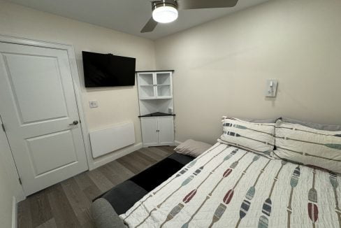 A neatly arranged, small bedroom with a bed, mounted television, and storage furniture.