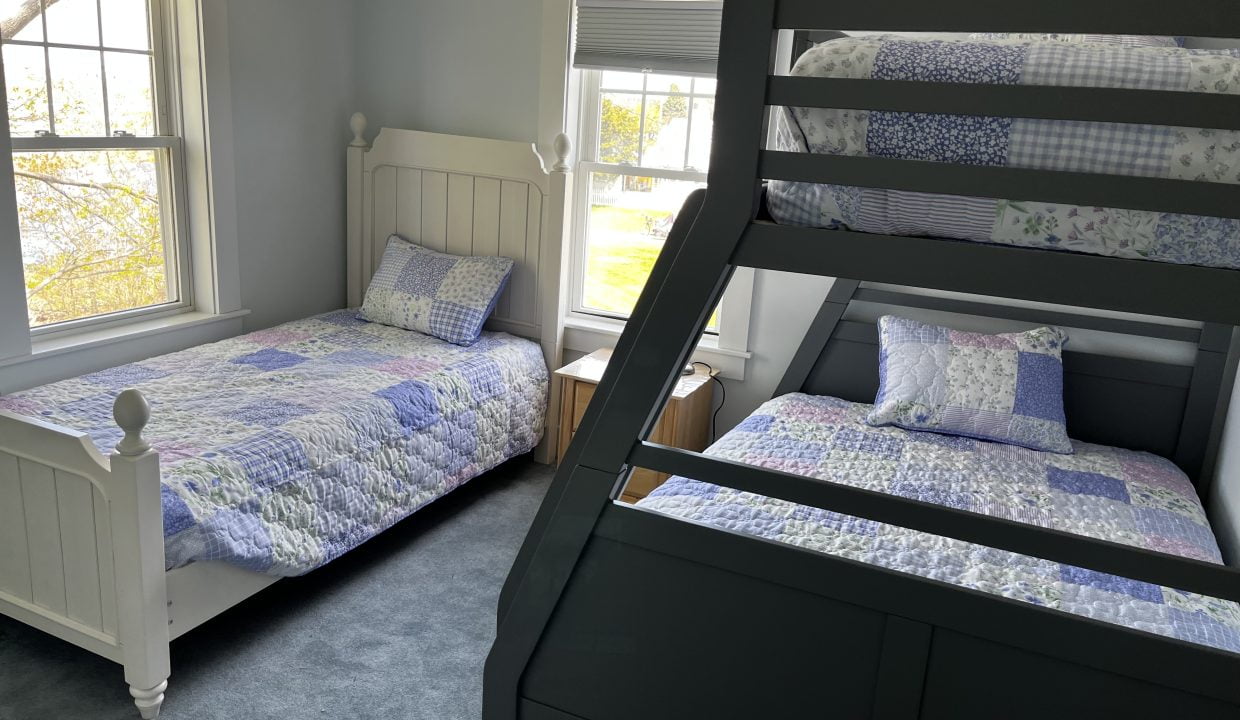 A bright bedroom with a white single bed and a black bunk bed, both covered with patchwork quilts, near windows showing greenery outside.