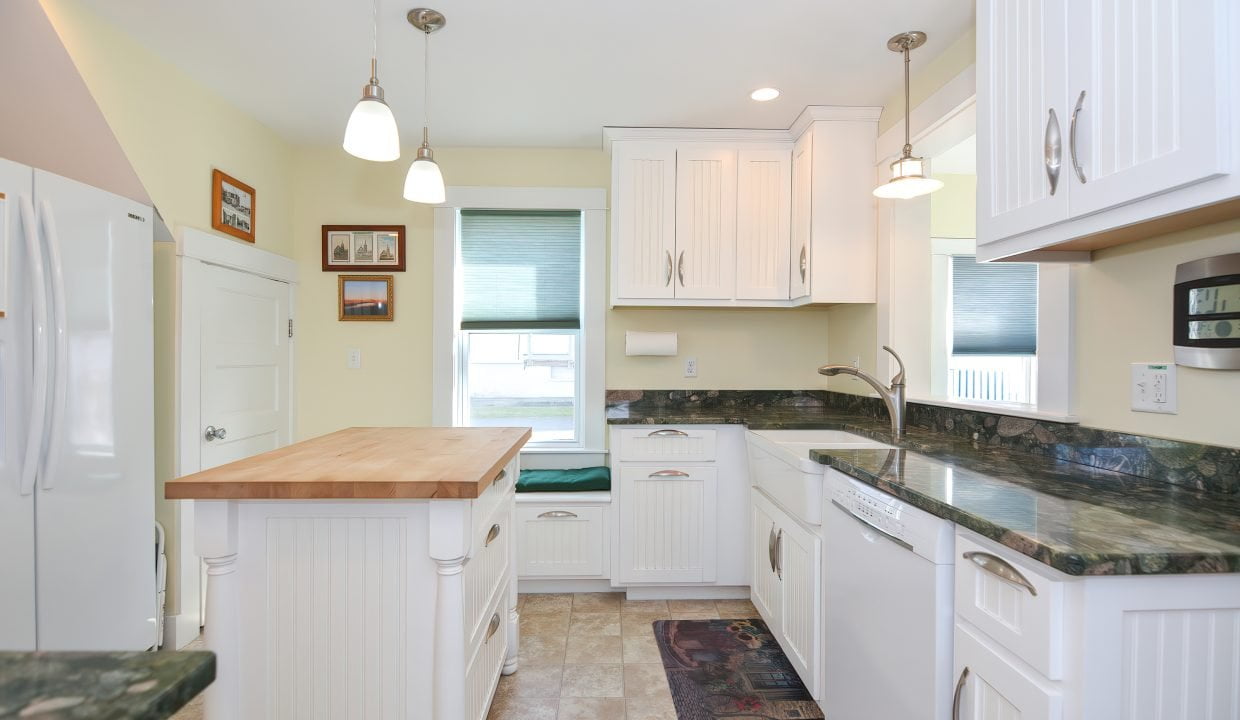 Bright kitchen with white cabinets, green marble countertops, pendant lights, and a wooden island, viewed from one end showing appliances and a window.