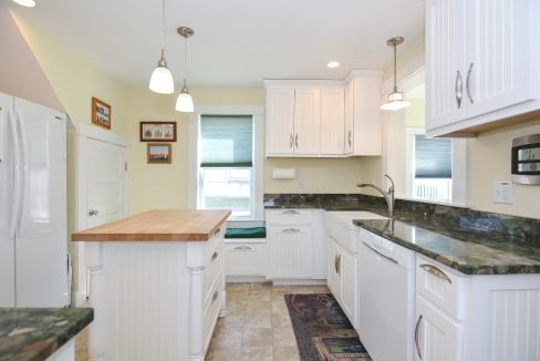 Bright kitchen with white cabinets, green marble countertops, pendant lights, and a wooden island, viewed from one end showing appliances and a window.