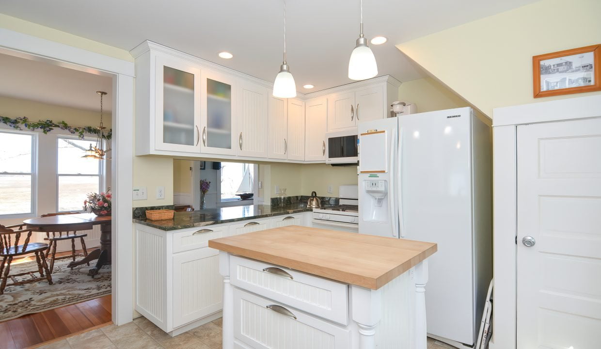 A bright, modern kitchen with white cabinetry, stainless steel appliances, a central wooden island, pendant lights, and a dining area visible in the background.