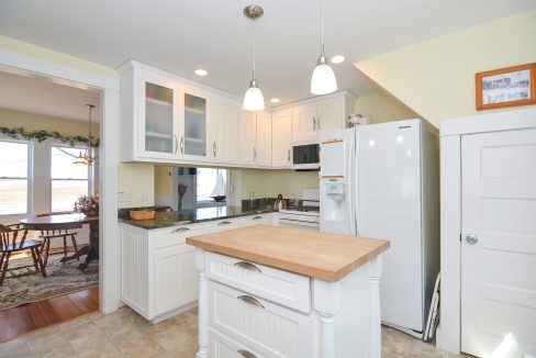 A bright, modern kitchen with white cabinetry, stainless steel appliances, a central wooden island, pendant lights, and a dining area visible in the background.