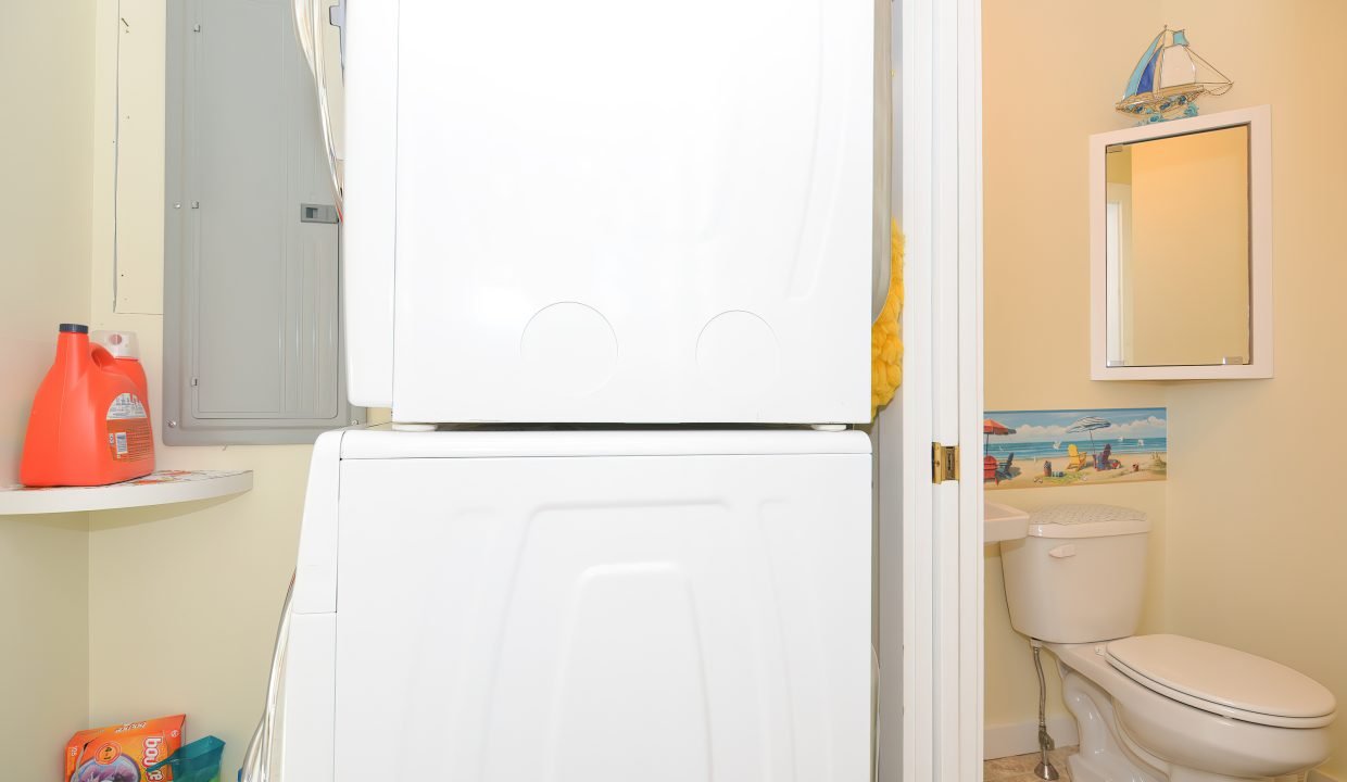 A small bathroom with a stacked washer and dryer, a toilet, and a mirrored cabinet. cleaning supplies are visible.