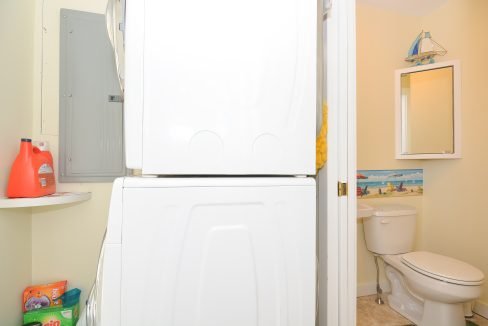 A small bathroom with a stacked washer and dryer, a toilet, and a mirrored cabinet. cleaning supplies are visible.