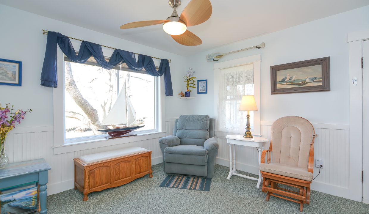 A cozy sunlit living room with a recliner, rocking chair, ceiling fan, and a window overlooking a tree. decor includes a floor lamp and nautical-themed paintings.