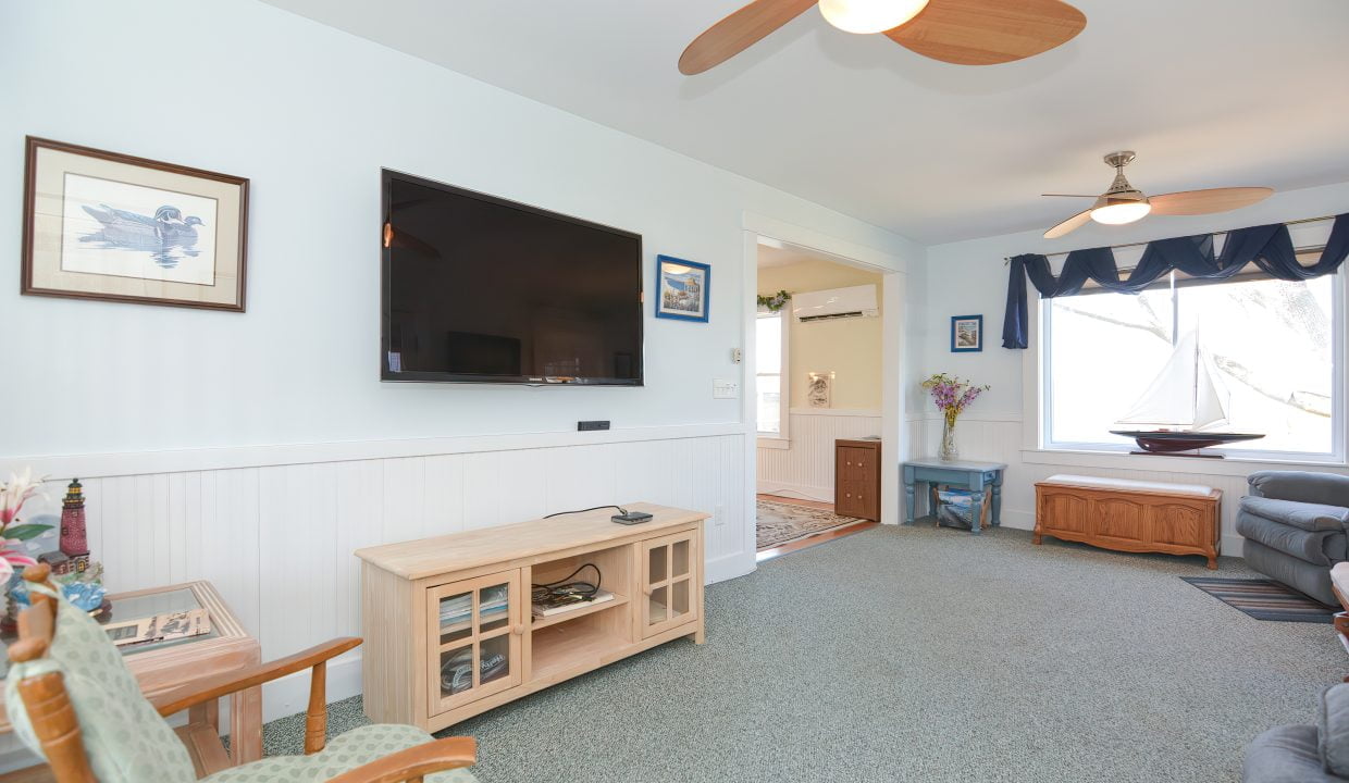 A bright living room with white and blue walls, a wall-mounted tv, a wooden tv stand, a ceiling fan, and a vertical ship-themed artwork.