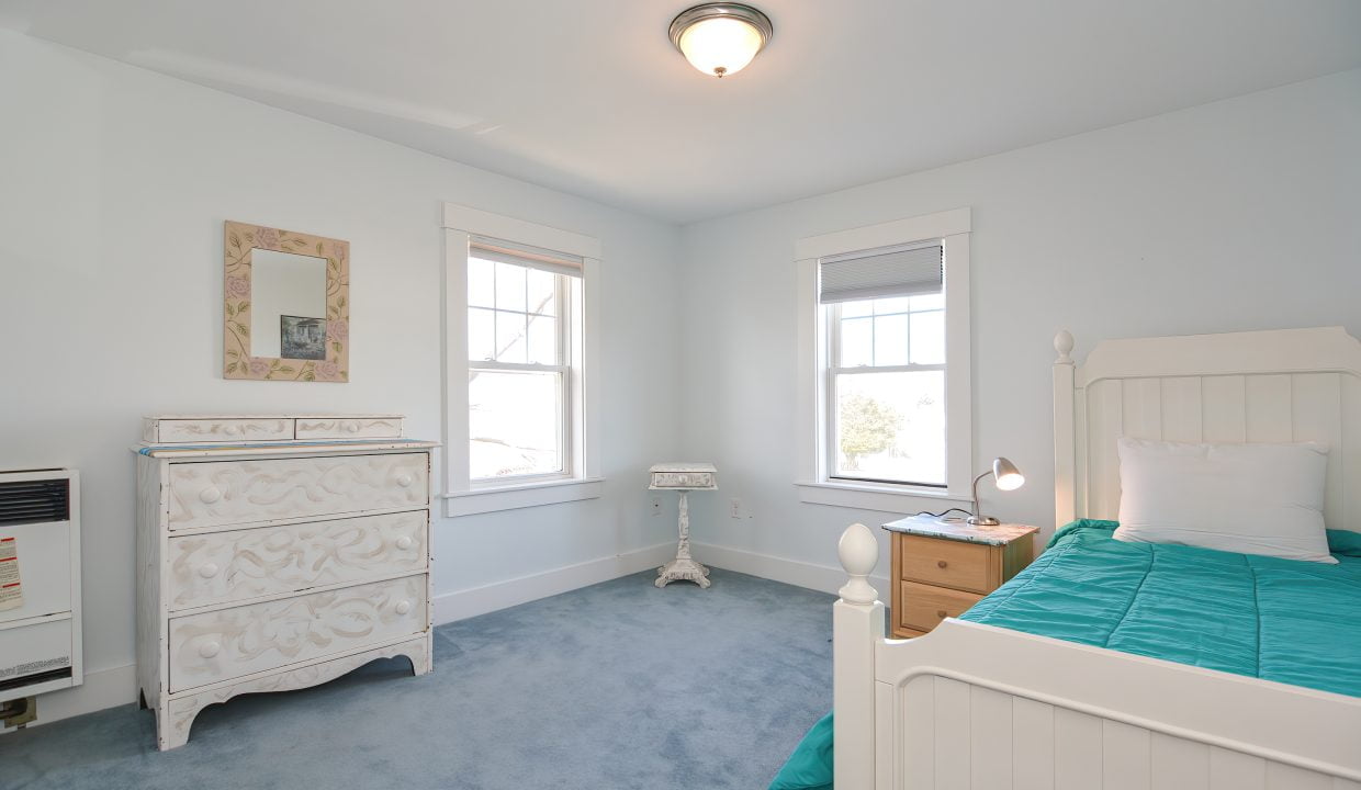Bright bedroom with blue carpet, white dresser, and bed with a teal bedspread, featuring two windows and a ceiling light.