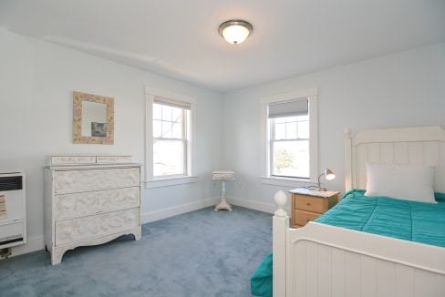 Bright bedroom with blue carpet, white dresser, and bed with a teal bedspread, featuring two windows and a ceiling light.