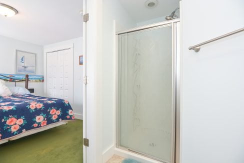 View from a bathroom showing an open door leading to a bedroom with a blue bedspread and floral accents, featuring a glass shower door in the foreground.