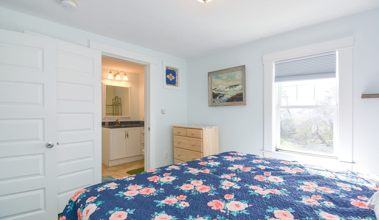 A bright bedroom with a floral blue bedspread, a wooden dresser, white doors, and a painting above the bed, leading to an en-suite bathroom.