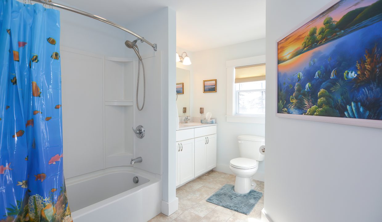 Bright bathroom with blue walls featuring a shower/bathtub with a colorful ocean-themed curtain, a toilet, white vanity, and ocean artwork.