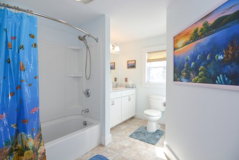 Bright bathroom with blue walls featuring a shower/bathtub with a colorful ocean-themed curtain, a toilet, white vanity, and ocean artwork.