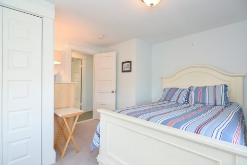 A small, neatly arranged bedroom with a white bed covered in blue and red striped bedding, an open door to a bathroom, and a wooden chair by a desk.