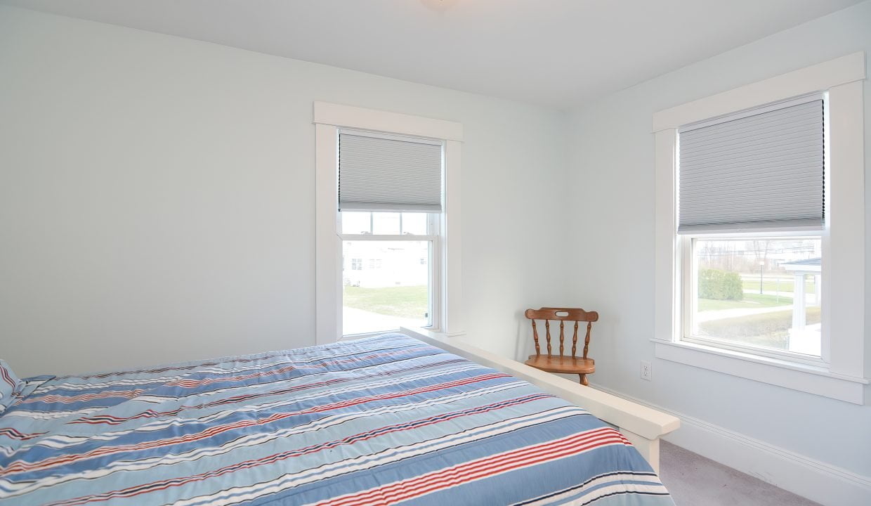 A simple bedroom with a bed covered in a striped blue and red comforter, a wooden chair by a window with white blinds, and white walls.