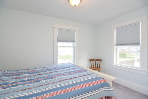 A simple bedroom with a bed covered in a striped blue and red comforter, a wooden chair by a window with white blinds, and white walls.