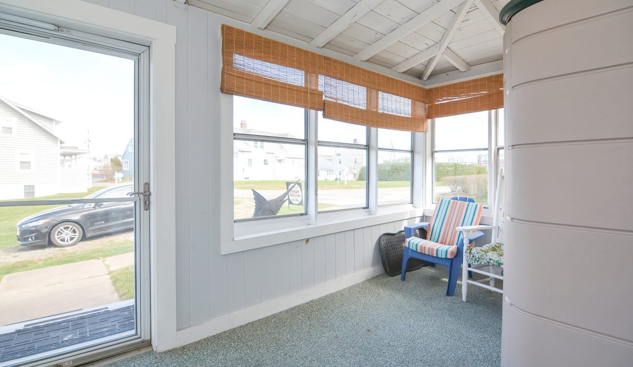 Bright, enclosed porch with wide windows, blue cushioned chairs, and bamboo blinds, overlooking a quiet residential street.