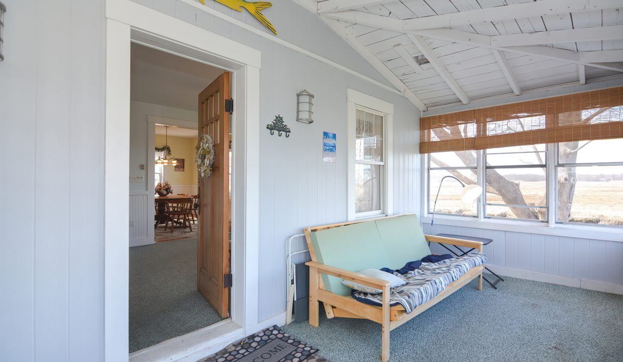 Bright sunroom with a light blue bench, a welcome mat, and wall decorations, looking into a cozy dining room in a coastal style house.