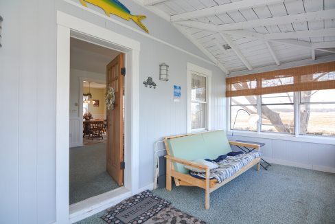 Bright sunroom with a light blue bench, a welcome mat, and wall decorations, looking into a cozy dining room in a coastal style house.