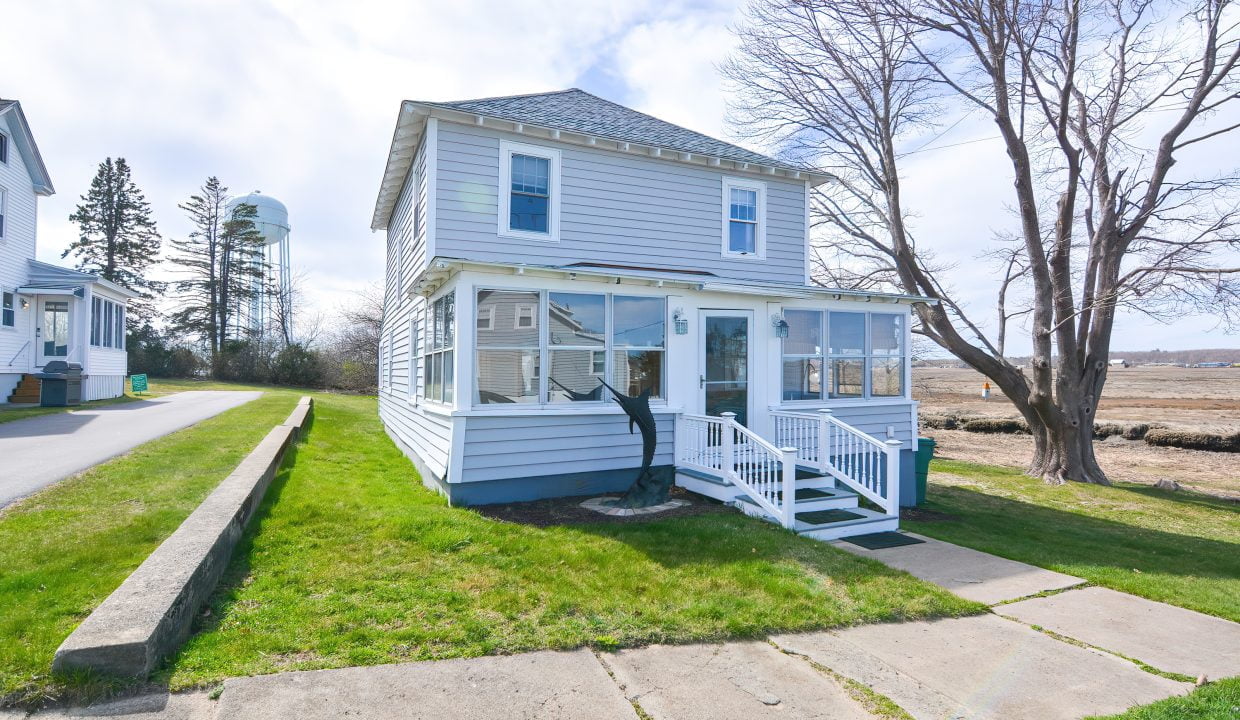 Two-story white house with blue trim and a front porch near a water body, with a clear sky and a water tower in the background.