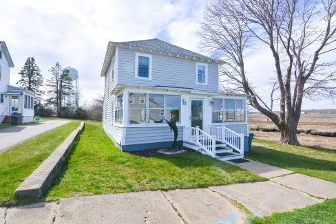 Two-story white house with blue trim and a front porch near a water body, with a clear sky and a water tower in the background.