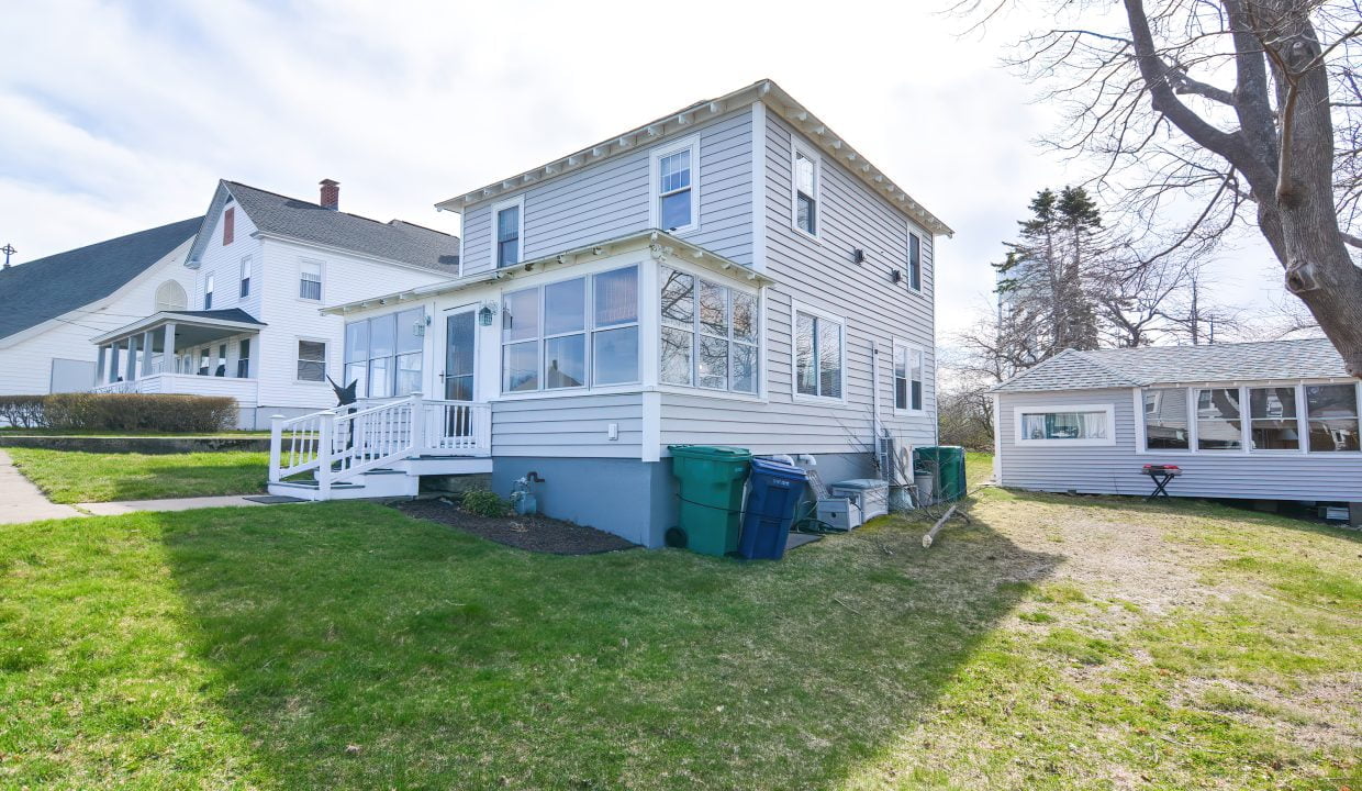 Two-story suburban home with a screened porch and detached garage, green lawn, and trash cans on the side under a clear sky.