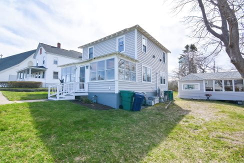 Two-story suburban home with a screened porch and detached garage, green lawn, and trash cans on the side under a clear sky.